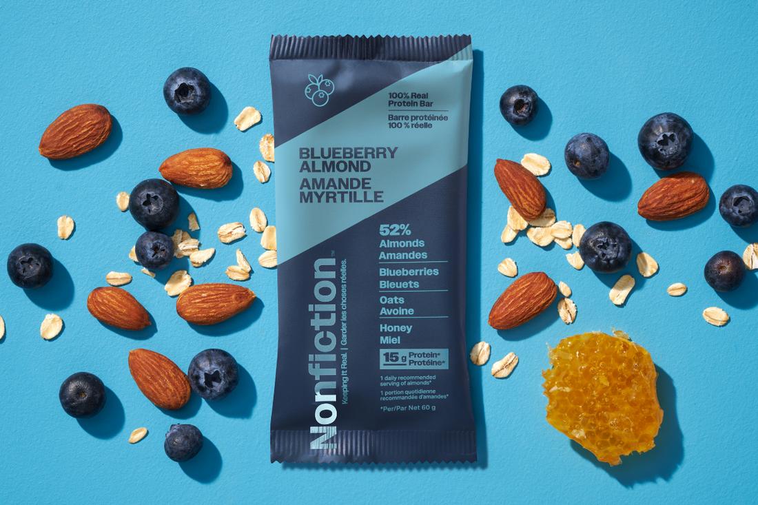 nonfiction blueberry almond protein bar on an orange background surrounded by bueberries, almonds, and raw honey.